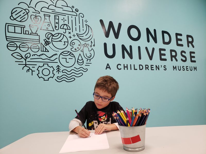 Picture of child drawing with colored pencil in front of Wonder Universe Logo. 