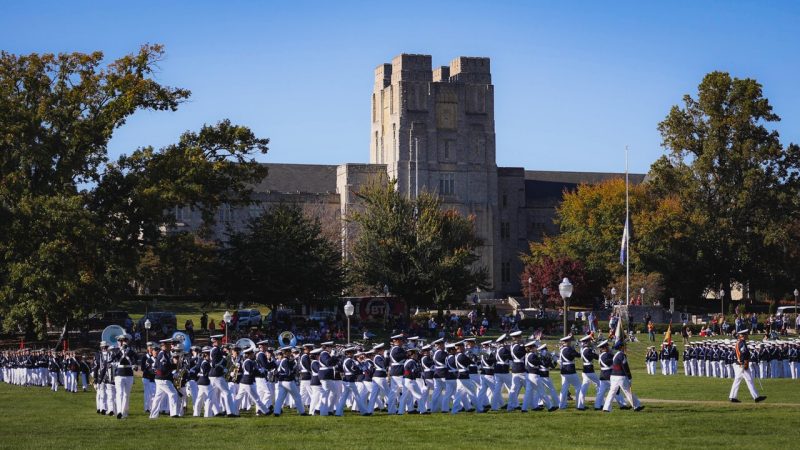 Cadets in white dress pants and navy jackets march in formation in front of Burruss Hall 